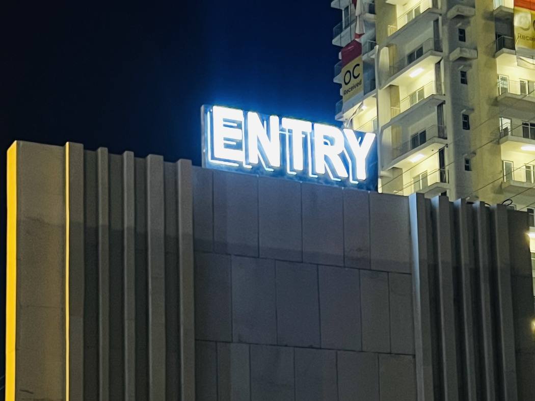 Lit lettering standing clear on top of a fluted stone-clad entrance gate, sized to be read from the approach road rather than from the gate itself.