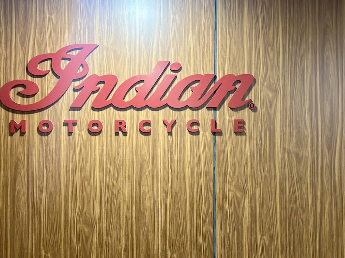 A script mark and block lettering cut as separate pieces and mounted on a wood-grain panelled wall, with a slim vertical light line run down the panel joint beside them.