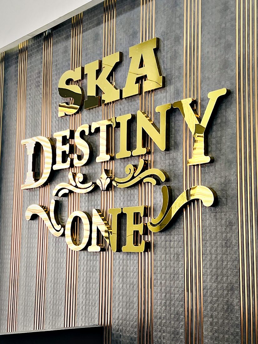 Ornate dimensional lettering in a gold metal finish, mounted on a fluted metal backdrop at the project entrance.
