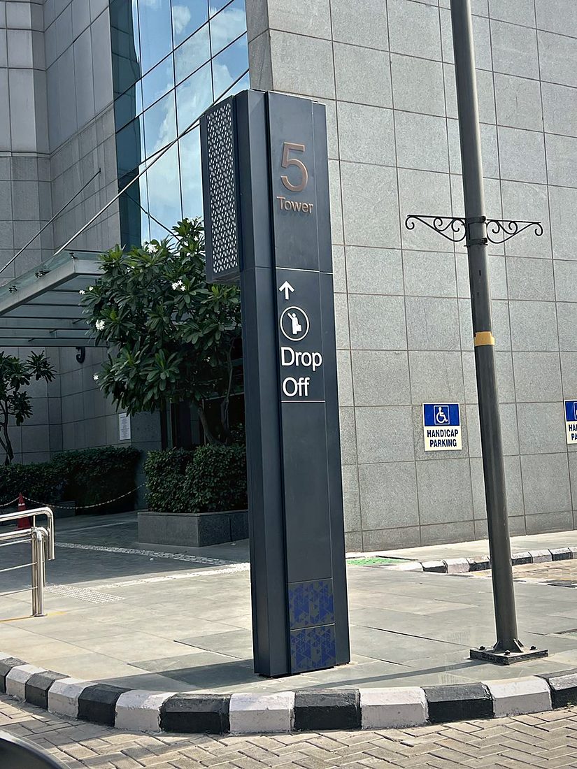 Slim tower-identification monolith in powder-coated steel with bronze-finish numerals and a perforated detail panel, marking the drop-off point at a corporate tower entrance.