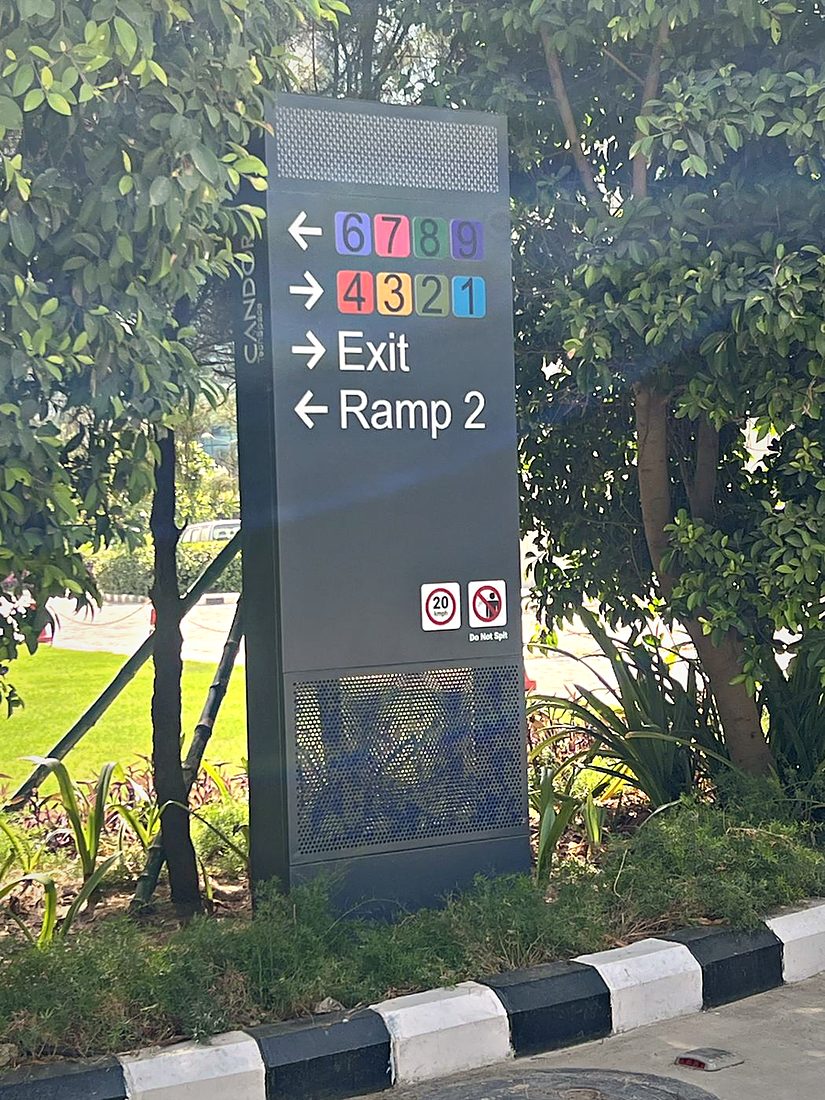 Vehicle-side directional totem using colour-coded tower numbers, exit and ramp arrows and speed pictograms to route drivers across a large basement parking layout.