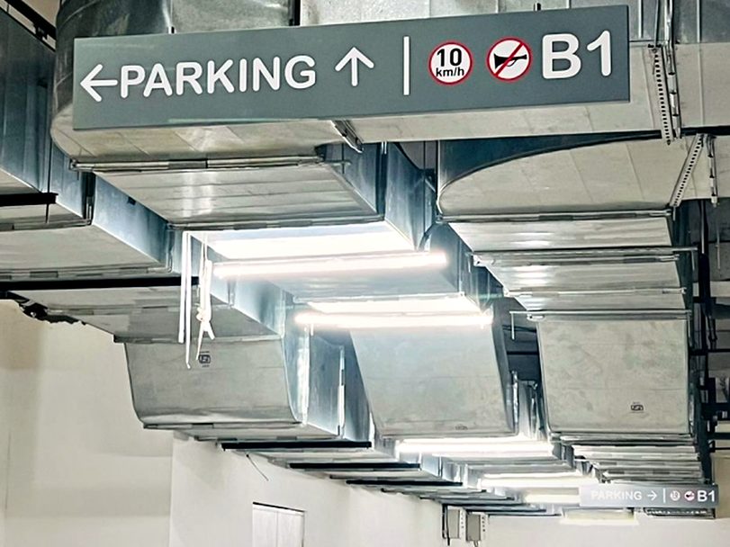 Suspended directional blade in a basement car park combining parking direction, level identification, speed limit and no-honking pictograms on one panel.
